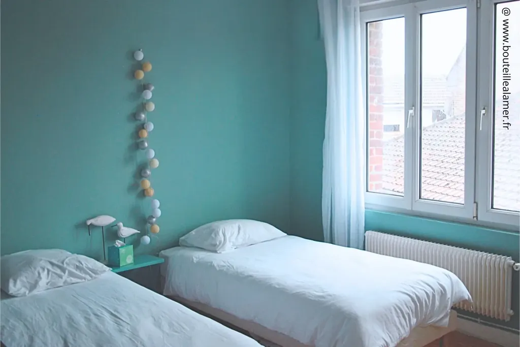 Chambre lumineuse avec deux lits simples, murs bleu-vert, grande fenêtre donnant sur le jardin côté bourg, gîte Bouteille à la Mer, Côte d’Opale, Gravelines, maison de vacances en location.