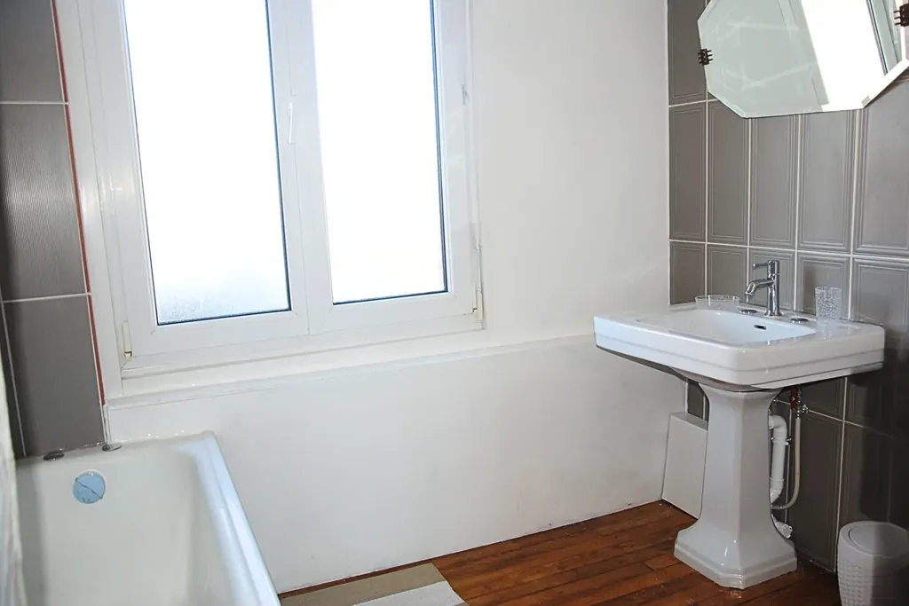 Salle de bain lumineuse au 2ᵉ étage avec baignoire, lavabo sur colonne, fenêtre à vitrage dépoli, sol en bois et carrelage gris, gîte Bouteille à la Mer, Côte d’Opale, Gravelines.