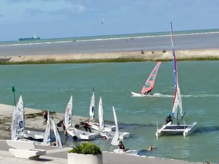 Vue panoramique depuis la chambre côté embouchure de Bouteille A la Mer® face à la mer et Gravelines : voiliers, windsurfers sur l'embouchure de l'AA, plage, et passage de bateaux en mer du Nord, Côte d'Opale.