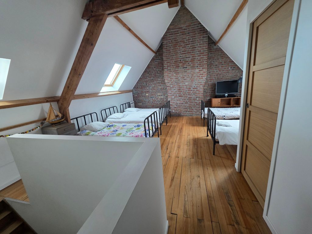 Dortoir mansardé avec poutres apparentes, mur en briques et cinq lits simples en fer forgé. Une vaste chambre idéale pour de belles vacances famille littoral Nord.