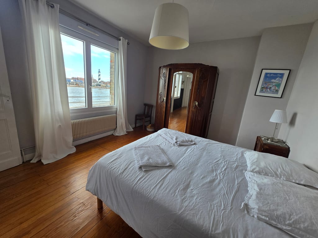 Chambre double lumineuse avec parquet ancien, armoire en bois et fenêtre offrant une vue directe sur le phare, idéale pour une location saisonnière à Grand-Fort-Philippe.