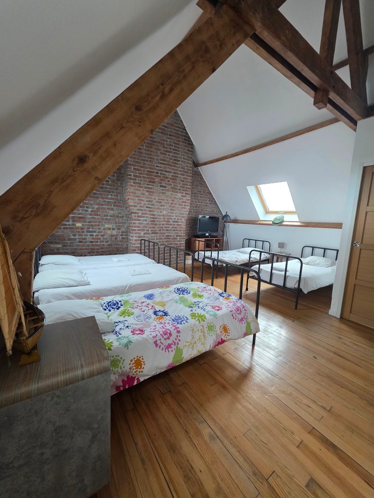 Vaste dortoir mansardé avec poutres apparentes en bois, mur de briques et lits simples. Une grande chambre familiale idéale pour les enfants dans notre gîte 4 étoiles à Gravelines.