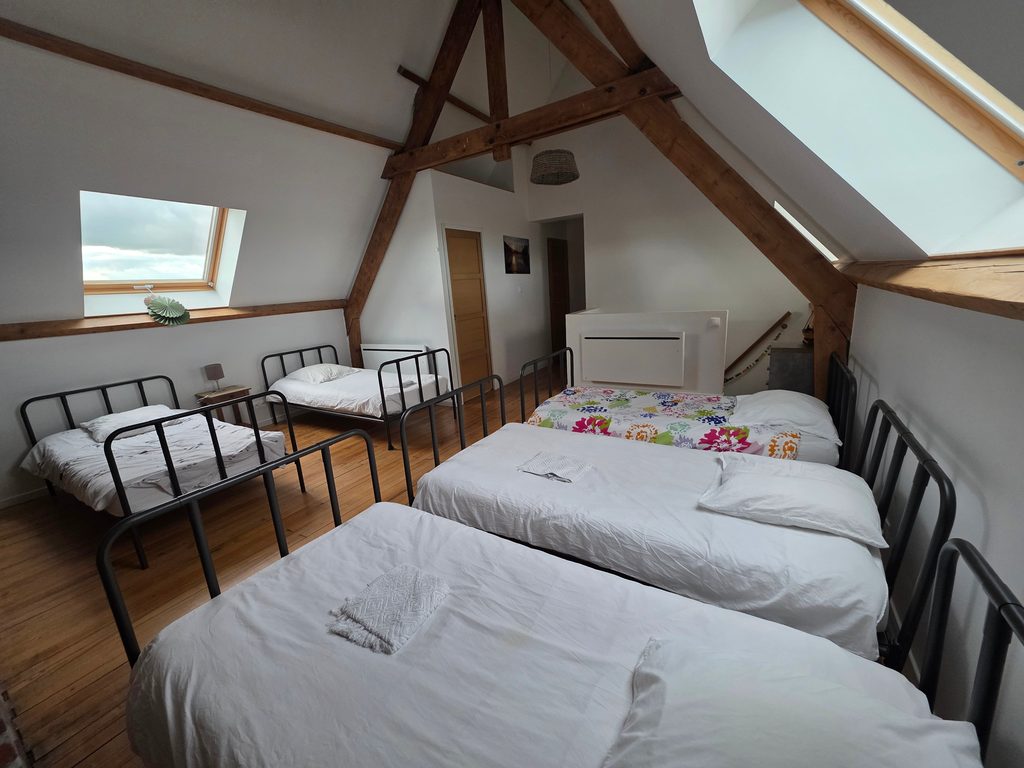 Vaste dortoir mansardé avec poutres apparentes en bois, mur de briques et lits simples. Une grande chambre familiale idéale pour les enfants dans notre gîte 4 étoiles à Gravelines.