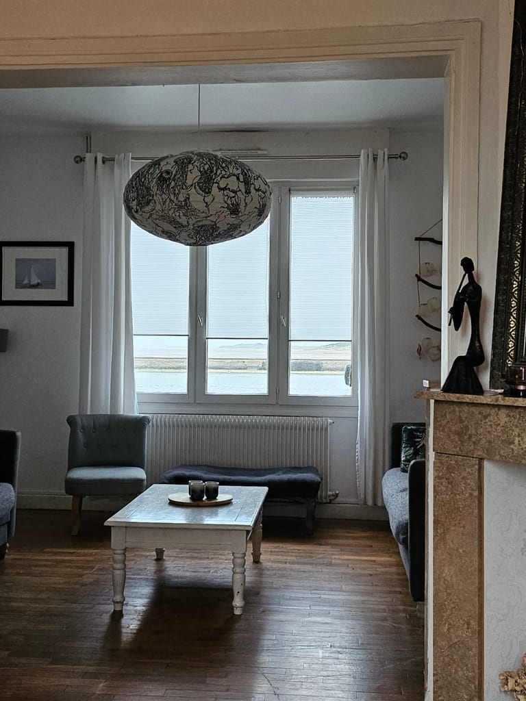Salon lumineux avec parquet en bois, canapés confortables et grande fenêtre offrant une vue dégagée sur l'eau. Cet hébergement Dunkerque bord de mer propose une décoration marine authentique et chaleureuse.