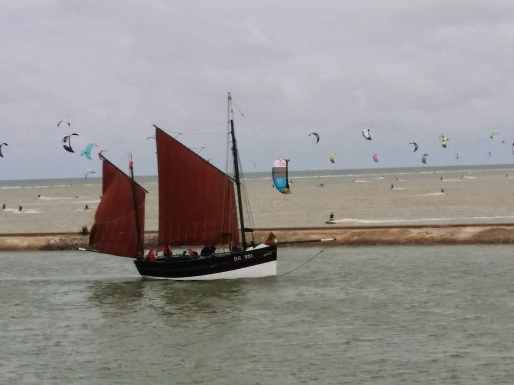 Vue depuis le premier étage du gîte sur un Dundee aux voiles brunes naviguant sur le chenal, avec des kitesurfeurs en arrière-plan. Une vue typique de notre location vacances Côte d'Opale.
